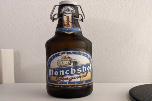 Eine Flasche Münchner Bier mit einem Aufkleber auf einem weißen Tisch, neben einem schwarzen Objekt, vor einer weißen Wand.