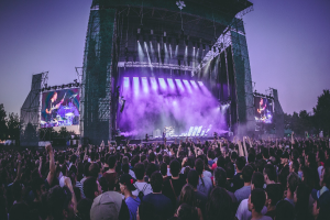 Menschenmenge auf einem Musikfestival mit Künstlern auf der Bühne, beleuchtet von Lichtern und umgeben von Bildschirmen, unter einem klaren blauen Himmel mit Bäumen und einem Gebäude im Hintergrund.