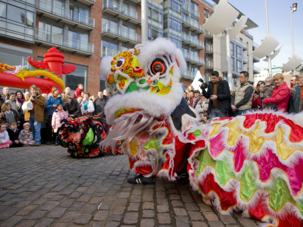 Ein lebendiges chinesisches Neujahrsfest in Amsterdam mit einem Löwen tanzen und einer Menge Menschen, einige halten Kameras, vor einem Hintergrund aus Gebäuden, Laternenmasten und einem klaren blauen Himmel.