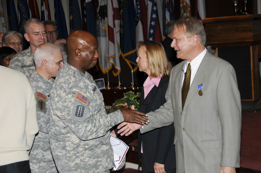 Eine Gruppe von Menschen, die mit zwei Personen Handsch├╝tteln, Flaggen im Hintergrund, ein Podium mit Mikrofon und eine Topfpflanze in der N├Ąhe.