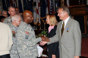 Eine Gruppe von Menschen, die mit zwei Personen Handsch├╝tteln, Flaggen im Hintergrund, ein Podium mit Mikrofon und eine Topfpflanze in der N├Ąhe.