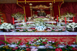 Idole auf einem Tisch mit Blumenvasen und Kerzenständern, die Kerzen halten, mit Blumen und Blättern unten und Vorhängen im Hintergrund.