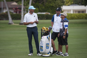 Mannergruppe auf einem grünen Feld mit einem Golfbag im Vordergrund, Bäume und Gebäude im Hintergrund.