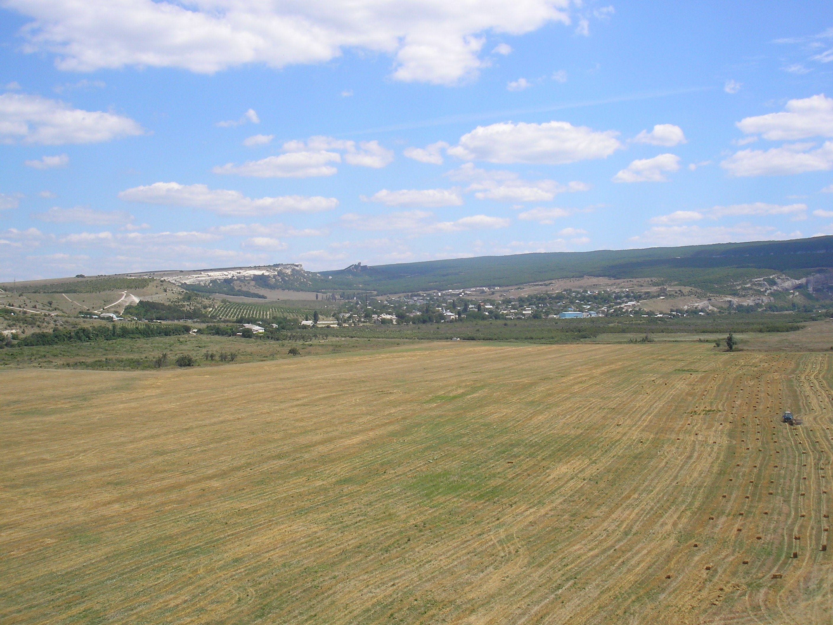 Ein Traktor arbeitet auf einem kultivierten Feld umgeben von Bäumen, Gebäuden, Hügeln und einem bewölkten Himmel.
