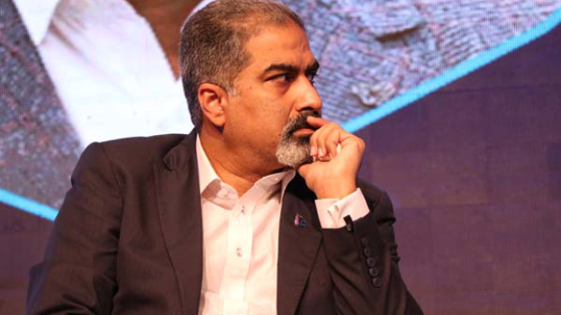 Ein Mann in Anzug und Krawatte sitzt auf einer Bühne und schaut nachdenklich auf einen Bildschirm hinter ihm.