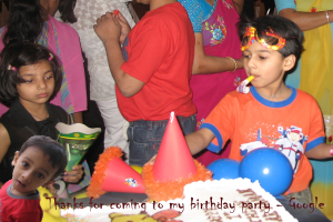 Kinder vor einem Tisch mit Kuchen, Luftballons und Mützen, mit anderen Menschen im Hintergrund.