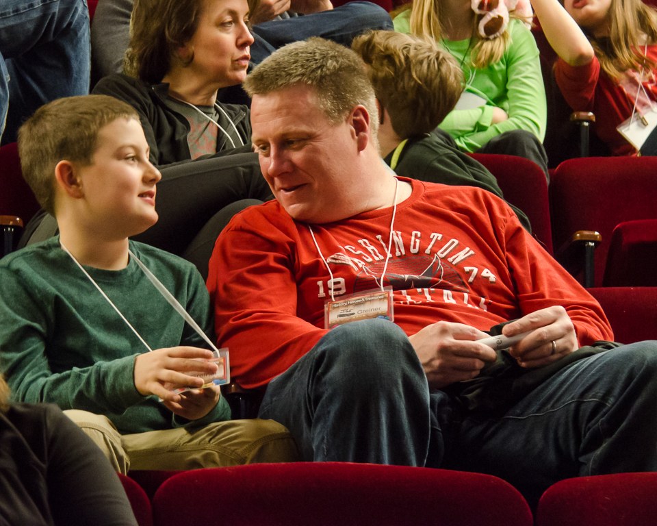 Eine Gruppe von Menschen sitzt in einem Kino, mit einem Mann und einem Jungen in der Mitte, die ein Objekt halten, umgeben von anderen auf Stühlen.