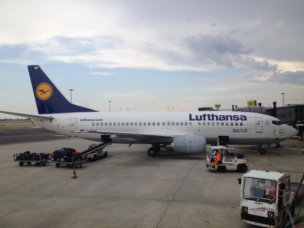 Ein Lufthansa-Airbus A320-200-Flugzeug steht auf dem Frankfurter Airport, umgeben von verschiedenen Bodenfahrzeugen, Wagen, Verkehrskegeln, Pfählen und Tafeln mit Text auf dem Rollfeld unter einem bewölkten Himmel.