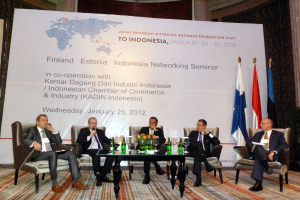 Eine Bühne mit Menschen, Stühlen, einem Mikrofon, Kabeln, einem Tisch, Flaschen, Blumen, Gläsern und anderen Gegenständen, mit Flaggen und einem Banner in der Mitte und einem Gebäude mit Vorhängen im Hintergrund.