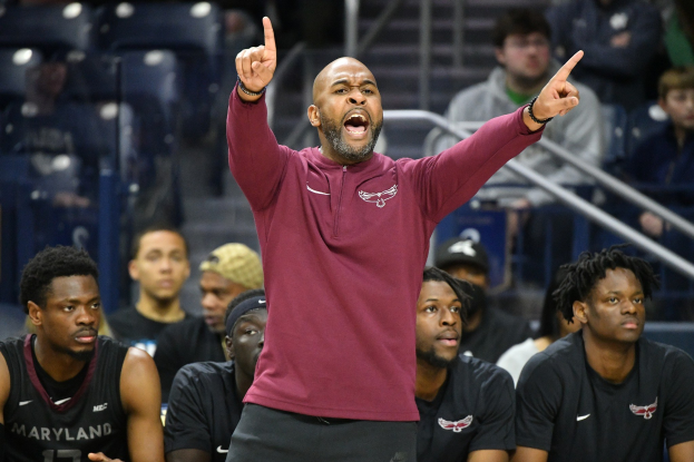 Cheftrainer des Maryland-Männer-Basketballteams in einem roten Hemd und schwarzen Shorts, der energisch gestikuliert, während er auf der Seitenlinie eines Basketballfelds steht, umgeben von sitzenden und stehenden Personen.