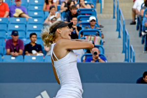 Eine Frau in weißer Oberbekleidung und schwarzer Mütze steht vor einem blauen Stadionplatz mit Zuschauern.