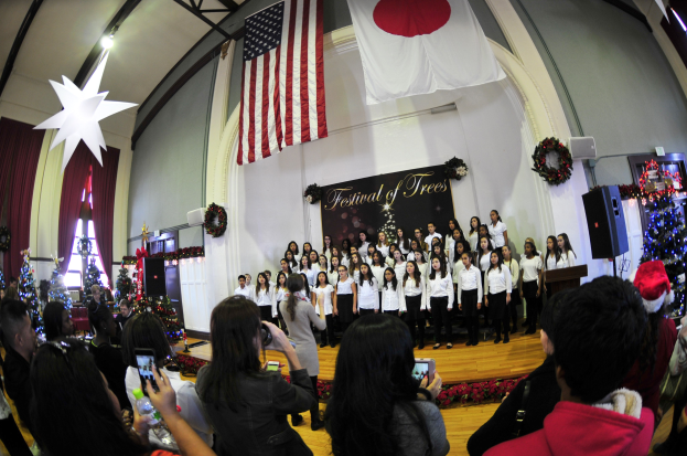 Eine Gruppe von Menschen auf einer Bühne mit Weihnachtsdekoration, einem Podium, Lautsprechern, einer Texttafel, Vorhängen, Fahnen und Deckenleuchten vor einem Publikum mit Kameras und Handys.
