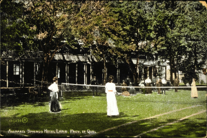 Schwarz-weißes Foto einer Gruppe von Menschen, die Tennis auf einem Rasenplatz vor dem Abenakis Springs Hotel in Provo, Quebec, spielen. Im Hintergrund sind Bäume und ein Gebäude zu sehen.