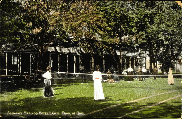 Schwarz-weißes Foto einer Gruppe von Menschen, die Tennis auf einem Rasenplatz vor dem Abenakis Springs Hotel in Provo, Quebec, spielen. Im Hintergrund sind Bäume und ein Gebäude zu sehen.