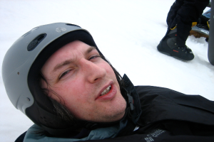 Ein Mann in einem Pullover und Helm steht im Schnee, mit den Beinen und Skiern einer anderen Person in der oberen rechten Ecke.