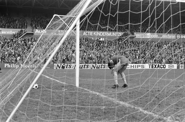 Schwarzes und weißes Foto eines Fußball-Torwarts bei einer Parade in der Nähe eines Torpfostens, mit einem Ball und Zuschauern im Hintergrund.