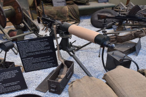 Eine Maschinengewehr auf einem Museumsausstellungsstück umgeben von verschiedenen Militärgeräten und Informationsschildern mit einer Wand im Hintergrund.