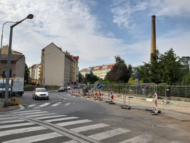 Stadtstraße mit parkenden Autos, Zebrastreifen, Verkehrskegel, Schilder, Laternen, Zaun, Bäume, Gebäude mit Fenstern und ein Schornstein unter einem bewölkten Himmel.
