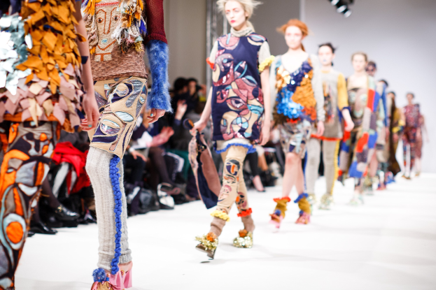 Eine Gruppe von Frauen, die auf dem London Fashion Week Laufsteg gehen, mit sitzenden Zuschauern und Bühnenbeleuchtung im Hintergrund.