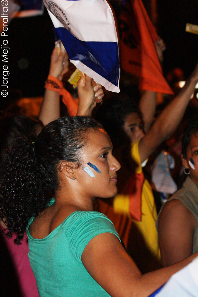 Eine Frau in einem gr√ľnen T-Shirt steht auf einer Demonstration und h√∂lt ein weißes Schild, mit vielen anderen Menschen hinter ihr.