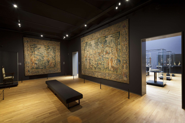 Eine Galerie im Museum of Fine Arts, New York, zeigt zahlreiche Gemälde an den Wänden, eine Bank, Pfosten mit Seilen, einen Stuhl, eine Tür, Deckenleuchten und verschiedene Gegenstände auf Tischen.