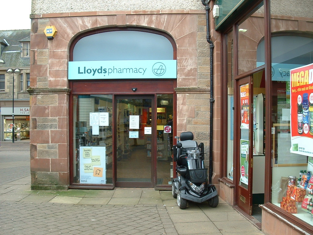 Eine Apotheke mit einem Fahrzeug davor und einem Gebäude links im Bild.