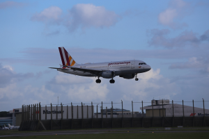 Germanwings Airbus A320-200 beim Start vom Frankfurt Airport mit Gras darunter, einem Zaun im Vordergrund, Gebäuden und Bäumen im Hintergrund und einem bewölkten Himmel darüber.