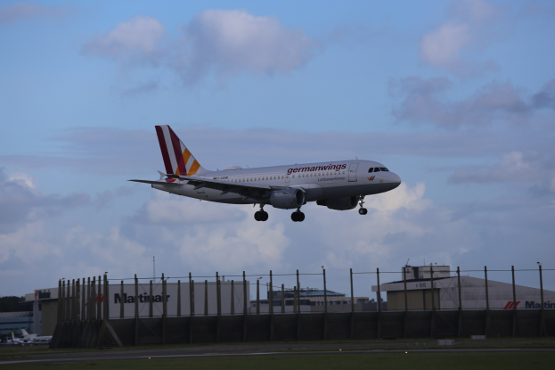 Germanwings Airbus A320-200 beim Start vom Frankfurt Airport mit Gras darunter, einem Zaun im Vordergrund, Gebäuden und Bäumen im Hintergrund und einem bewölkten Himmel darüber.