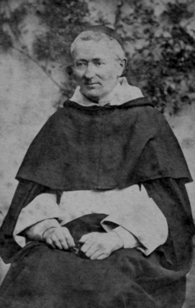 Altes Schwarz-Weiß-Foto eines Mannes in Priesterrobe, der vor Bäumen sitzt und eine schwarze Robe mit weißem Kragen und Manschetten trägt, die Hände zum Gebet gefaltet, das Gesicht von warmem Licht erhellt mit friedlichem Ausdruck.