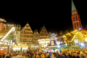 Ein lebendiger Weihnachtsmarkt in Nürnberg, Deutschland, mit Menschen um beleuchtete und dekorierte Stände, Gebäuden mit Fenstern und einem Kirchturm im Hintergrund unter einem dunklen Himmel.