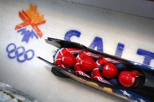 Ein Bobfahrer in einem Schlitten bei den Olympischen Spielen, mit einer dekorierten Wand und Text im Hintergrund.