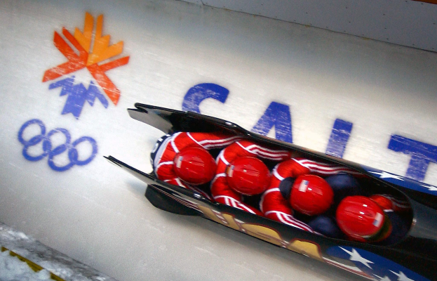 Ein Bobfahrer in einem Schlitten bei den Olympischen Spielen, mit einer dekorierten Wand und Text im Hintergrund.