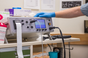 Eine Person in blauen Handschuhen arbeitet an einem medizinischen Gerät an einem unordentlichen Tisch in einer Krankenhausumgebung, mit Papieren an der Wand im Hintergrund.