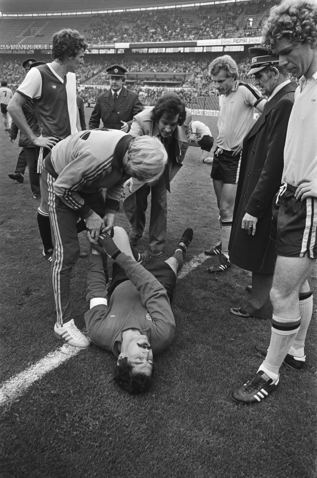 Schwarzes Bild eines verletzten Fussballspielers auf dem Boden umgeben von Teamkollegen und einem Stadionpublikum