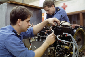 Zwei Männer in blauen Uniformen arbeiten an einem Flugzeugmotor in einer Fabrik, mit einem roten Kreuzsymbol im Hintergrund.