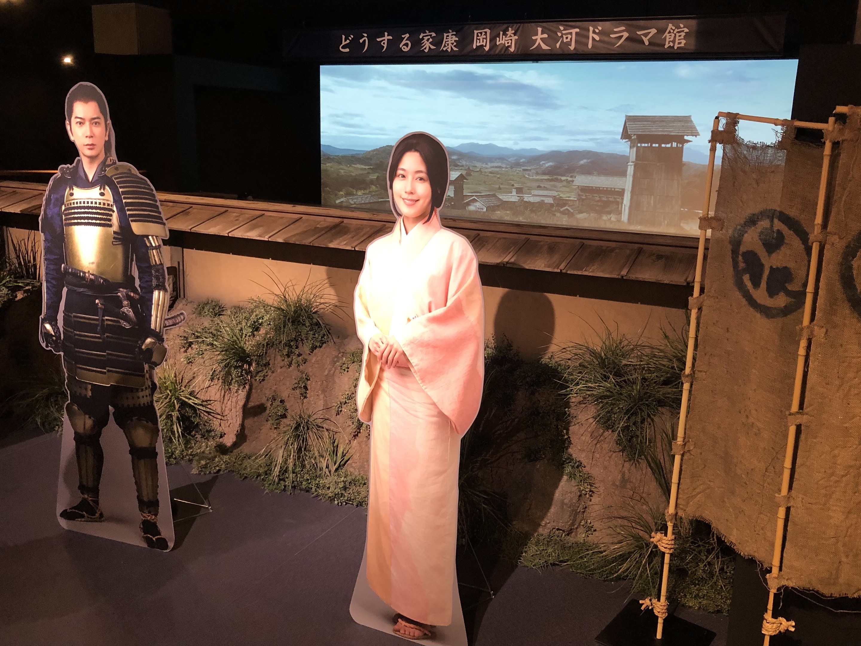 Zwei Personen - eine Frau in einem rosa Kimono und ein Mann in Samurai-Rüstung - lächeln in einer Szene aus einem japanischen Film, mit Pflanzen, einem Bildschirm und Beleuchtung im Hintergrund.