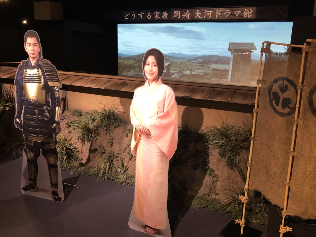Zwei Personen - eine Frau in einem rosa Kimono und ein Mann in Samurai-Rüstung - lächeln in einer Szene aus einem japanischen Film, mit Pflanzen, einem Bildschirm und Beleuchtung im Hintergrund.