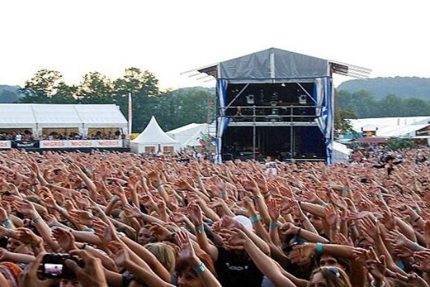 Eine große Menschenmenge auf einem Musikfestival mit erhobenen Händen, einige halten Handys, vor einer Bühne mit Künstlern, weißen Zelten, Bäumen und einem klaren blauen Himmel.