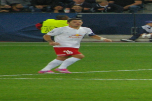Ein Mann in einem weißen Hemd und roten Shorts, der auf einem Feld Fußball spielt, mit einer Gruppe von Menschen im Hintergrund.