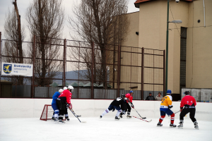 Menschen beim Eishockey auf einem Eisplatz mit Gebäuden, Bäumen und einer Straßenlaterne im Hintergrund.