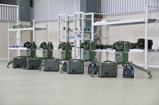 Gruppe von Militärrobotern auf einem Boden mit Koffern und Gegenständen auf Gestellen, in der Nähe einer Wand und eines Geländers als Teil eines Verteidigungssystems positioniert.