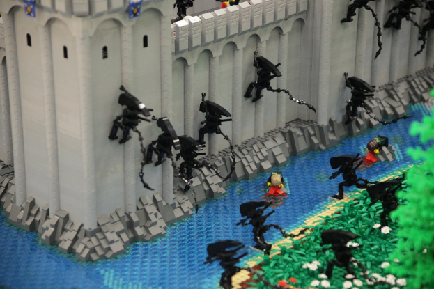 Ein Lego-Schlossmodell mit einer Gruppe von Menschen darauf, umgeben von verschiedenen Spielzeugen, Wasser, Pflanzen, Blumen und anderen Gegenständen.