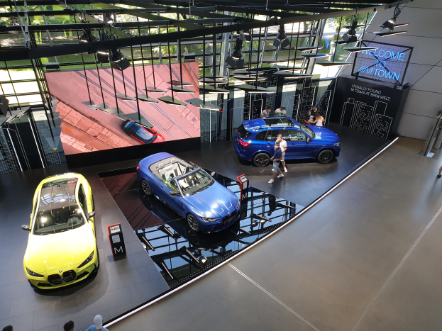 Eine Gruppe von Menschen steht im BMW Museum in München, Deutschland, mit Autos auf der linken Seite und einem Schild mit der Aufschrift "BMW Museum München" auf der rechten Seite, mit Blick auf Bäume, Gebäude und den Himmel durch Glasfenster.