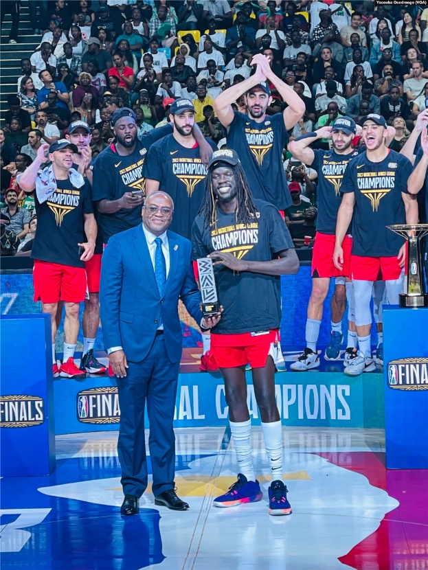 Gruppe von Männern auf einem Basketballfeld mit Trophäen, Zuschauern im Hintergrund und zwei Trophäen auf Podesten rechts.