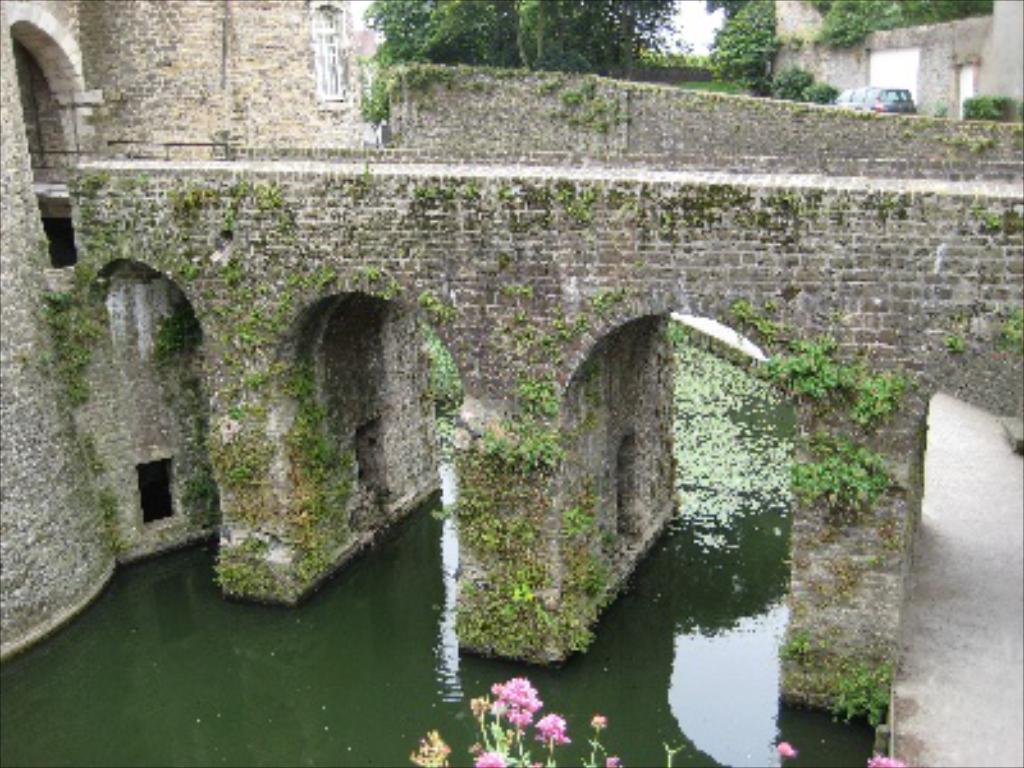 Eine Brücke in der Mitte des Bildes, mit einer Straße, einem Baum und Wasser unten, einem Auto und einer Wand oben rechts und einem Gebäude auf der linken Seite.