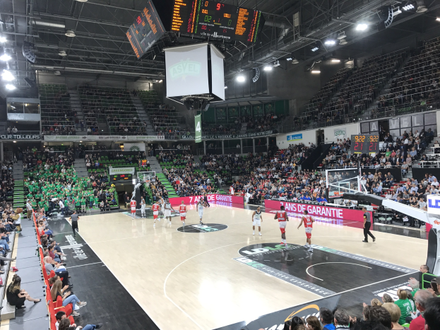 Basketballspiel in einer großen Arena mit Zuschauern, mit einem Spiel zwischen Asvel und Galatasaray, wie es auf den Texttafeln zu sehen ist.