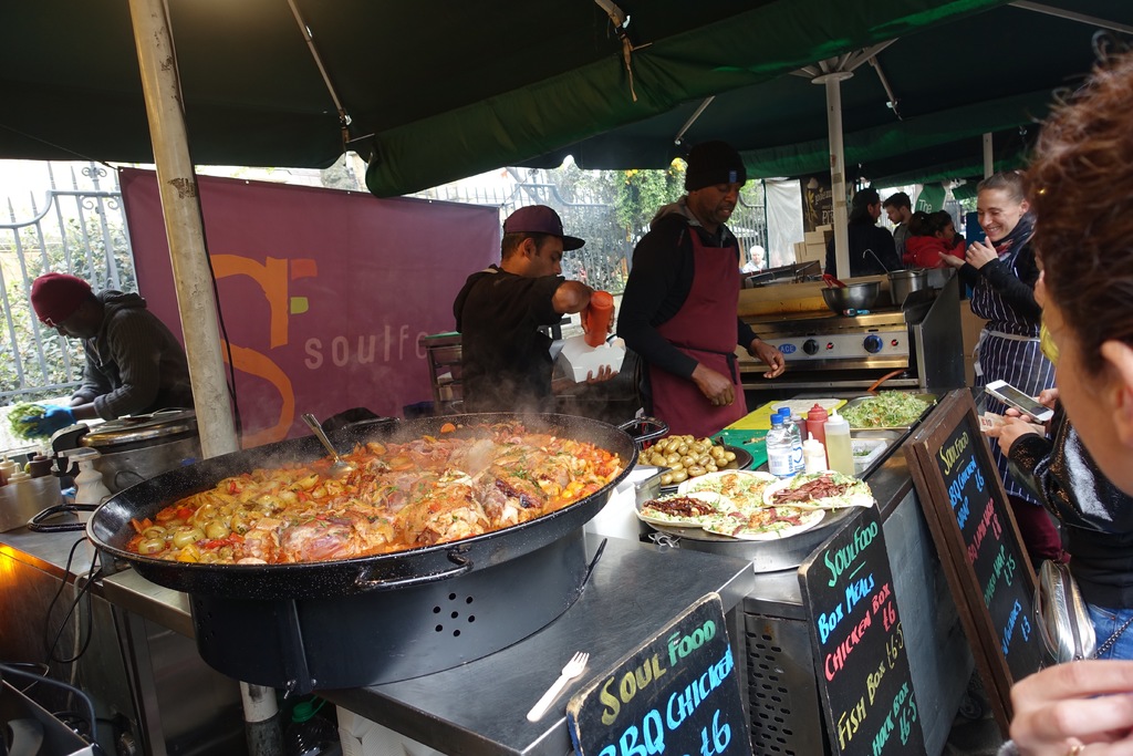 Ein Imbissstand mit einer Gruppe von Menschen, einer Person in der Mitte mit einer Flasche auf einer Kiste, einem Herd auf der rechten Seite, einer Tafel in der rechten unteren Ecke und Essen in der linken unteren Ecke.