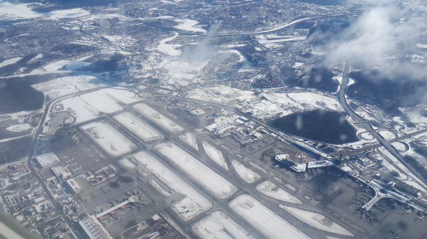 Luftaufnahme eines schneebedeckten Flughafens mit Gebäuden, Straßen, Brücken, Wasser, Bäumen und bewölktem Himmel.