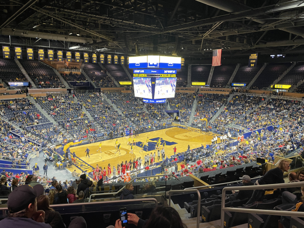 Eine große Arena mit Zuschauern, die ein Basketballspiel verfolgen, mit einer Person, die ein Mobiltelefon hält, Deckenleuchten und Schilder mit "Michigan Wolverines Basketball".