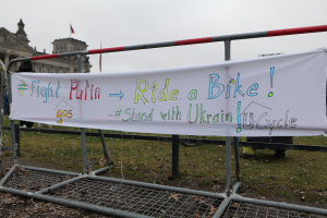 Eine Gruppe von Menschen hält ein Banner mit der Aufschrift "Kämpfe gegen Putin, fahr Rad, steh mit der ukrainischen U-Cycle" und steht auf dem Boden vor einem Metallzaun, trockenen Blättern, einem Gebäude mit Fenstern, einem Fahnenmast mit einer Flagge und einem bewölkten Himmel.